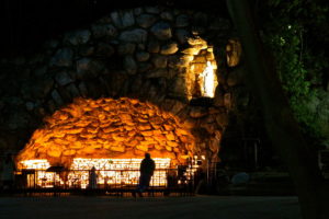 Picture was taken at night of the grotto of our lady lourdes university of notre dame, there's a person standing looking to another person on knees praying