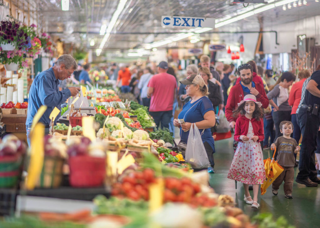 A view inside of South Bend Farmer's Market, a man is selling vegetables to a lady and there's people walking around